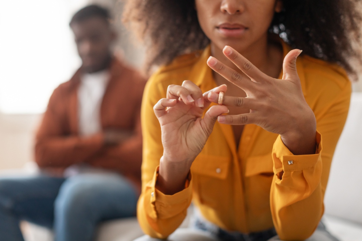 mujer quitándose el anillo de matrimonio, su marido en el fondo, de brazos cruzados