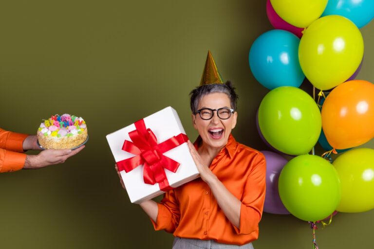mujer de mediana edad sosteniendo un regalo, unas manos acercándole un pastel y globos en el otro lado