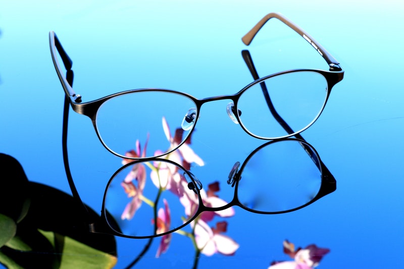 A pair of black framed clear glasses on a reflective bright blue top and an out of focus purple and green orchid flower.