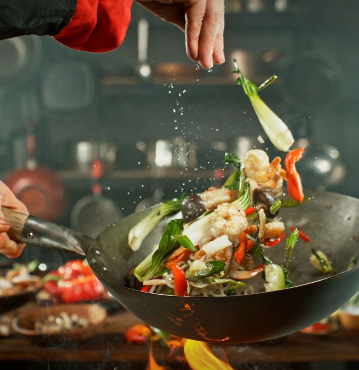 A deep frying pan tossing vegetables in the air and a hand adding salt.